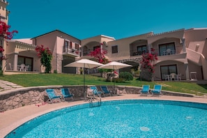 Piscina com espreguiçadeiras e guarda-sóis, rodeada por um edifício com varandas e uma parede de pedra.