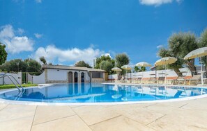 Ein Swimmingpool mit Liegestühlen und Sonnenschirmen, ein kleines Gebäude mit Bögen und Bäume im Hintergrund.