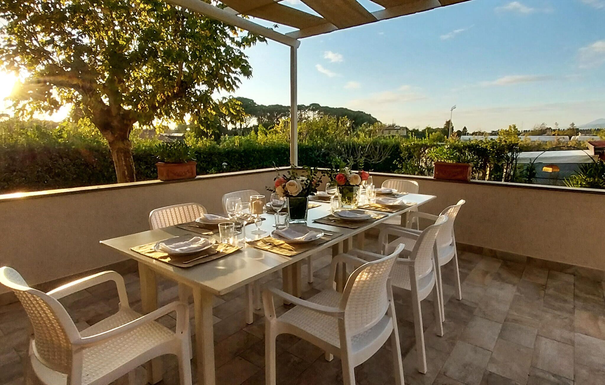 Un comedor con una mesa dispuesta para cuatro personas, rodeado de sillas blancas, bajo una pérgola con vistas a un jardín ajardinado y colinas lejanas.