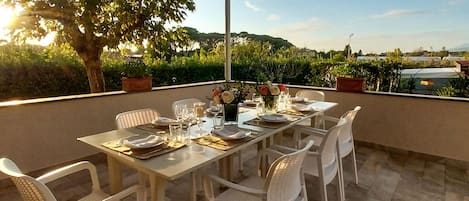 Un coin repas avec une table dressée pour quatre personnes, entourée de chaises blanches, sous une pergola d’où l’on peut admirer un jardin aménagé et des collines lointaines.