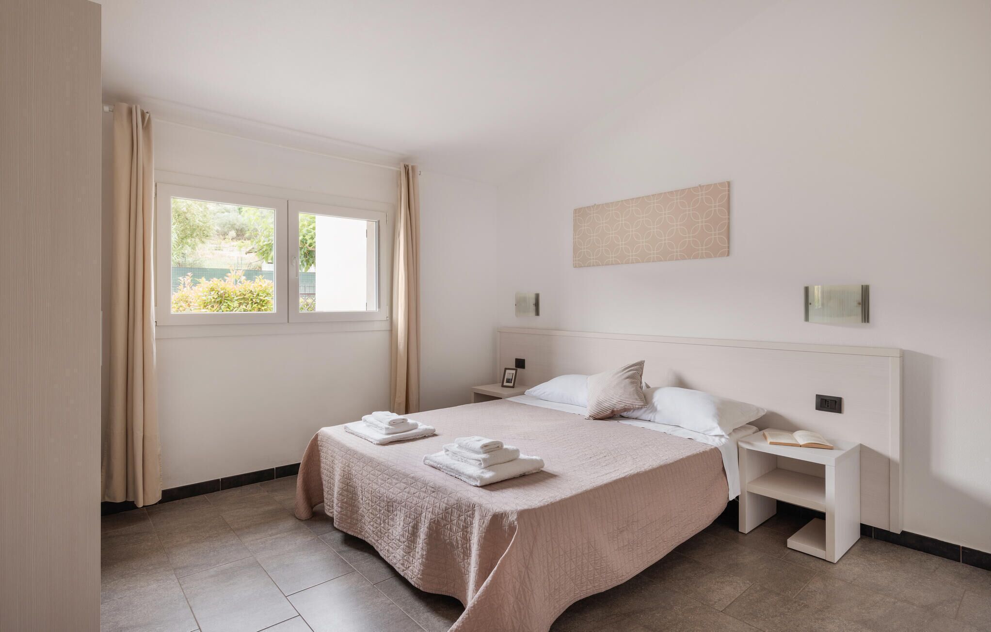 A hotel room with a large bed, a bedside table with a lamp, a window with curtains, and a view of greenery outside.