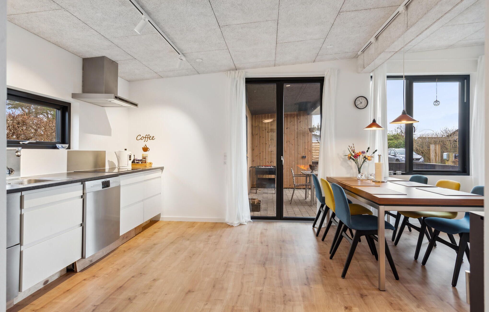 Eine moderne Küche mit weißen Schränken, einer Holz Arbeitsplatte und einer Kaffeestation. Es gibt einen Essbereich mit einem Holztisch und Stühlen, sowie einen Blick ins Freie durch eine Glastür.