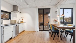 Eine moderne Küche mit weißen Schränken, einer Holz Arbeitsplatte und einer Kaffeestation. Es gibt einen Essbereich mit einem Holztisch und Stühlen, sowie einen Blick ins Freie durch eine Glastür.
