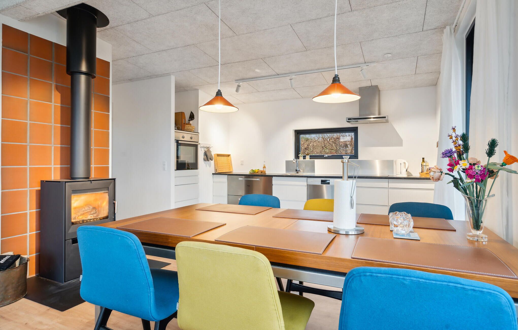 Una cocina moderna con estufa integrada, un comedor con mesa de madera y sillas coloridas, iluminación colgante y vistas al exterior a través de una ventana.