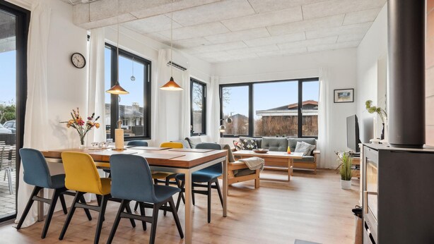 Ein modernes Wohnzimmer mit einem Holzesstisch, bunten Stühlen, einer Couch und einem großen Fenster mit Blick ins Freie.