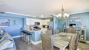 A modern kitchen with white cabinets, a central island, and a dining area with wicker chairs and a glass table. - Renovated oceanfront penthouse condo - direct beach access!  (Fort Walton Beach)