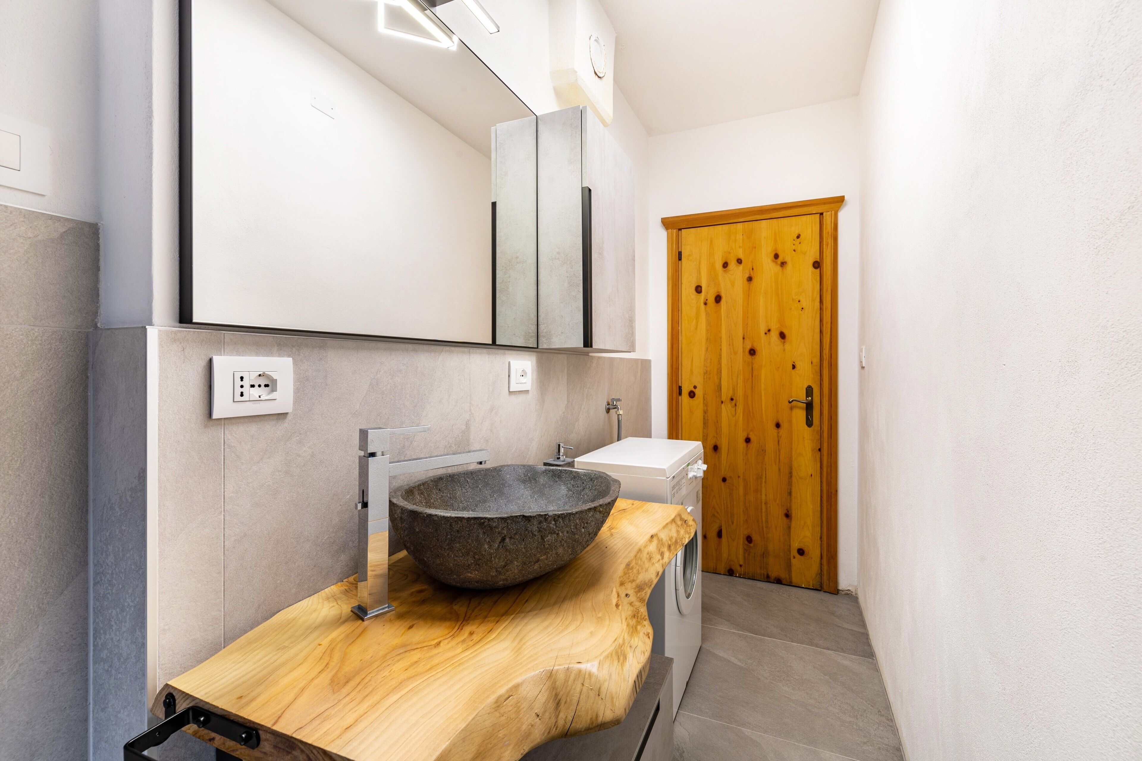 Un baño moderno con lavamanos de piedra, encimera de madera, espejo, lavadora y puerta de madera.