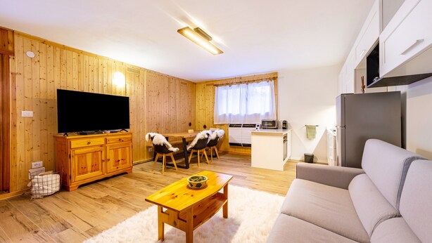 Una sala de estar con televisor, armarios de madera, una mesa de comedor con sillas, un sofá y una zona de cocina con refrigerador.
