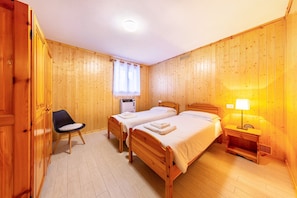 Una habitación con dos camas de madera, una silla, una mesita de noche con lámpara y una ventana con cortinas.