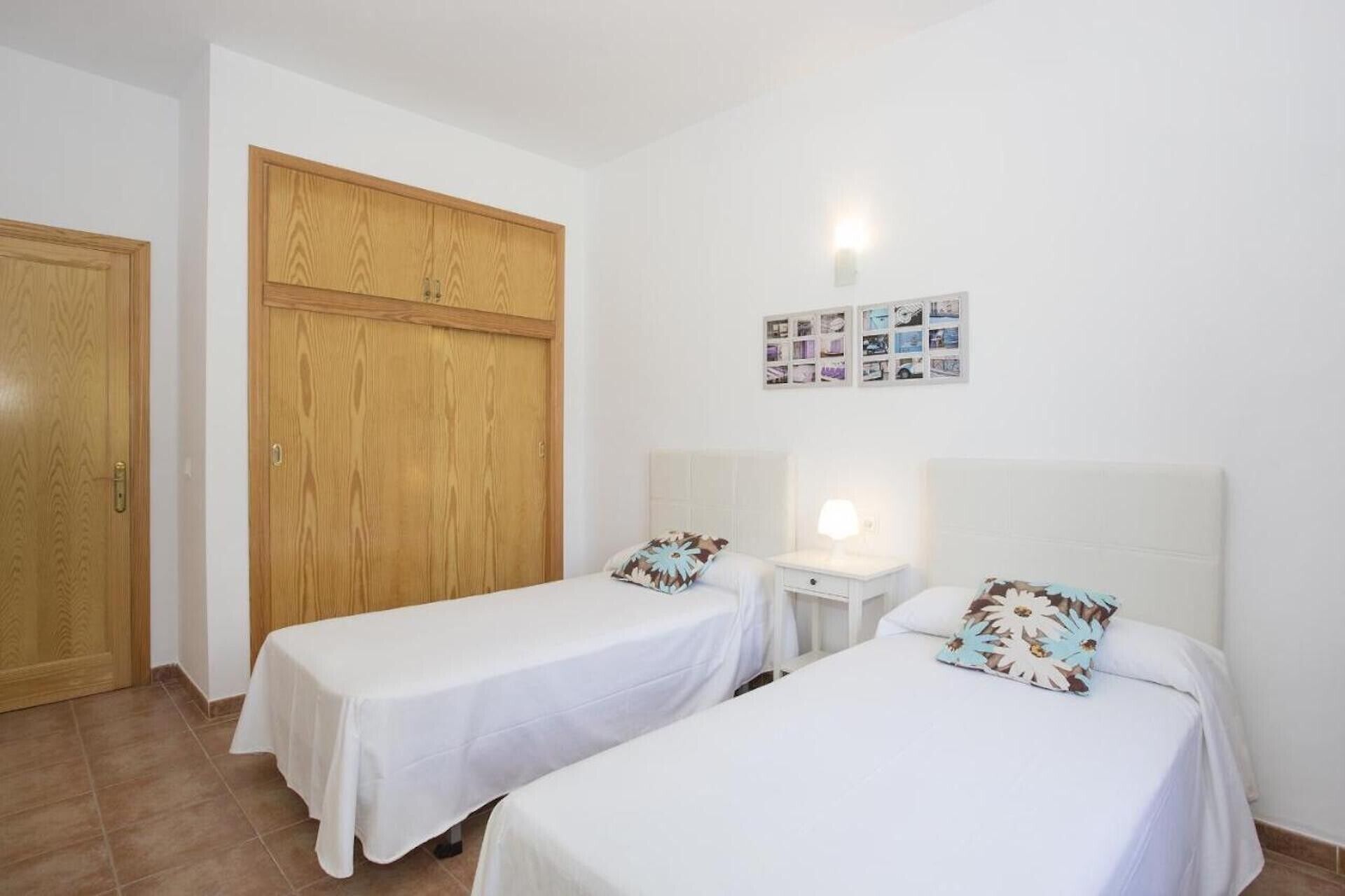 Ein Zimmer mit zwei Betten, einem Holzschrank, einem kleinen Nachttisch mit Lampe und einer Wand mit Fotos.