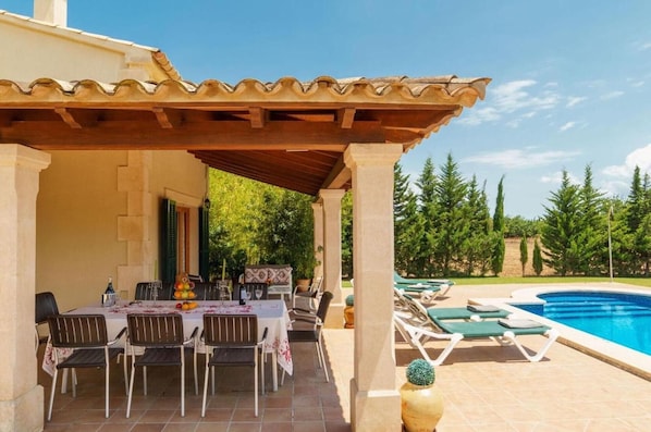 Un espacio comedor al aire libre techado con piscina y sillones de descanso.