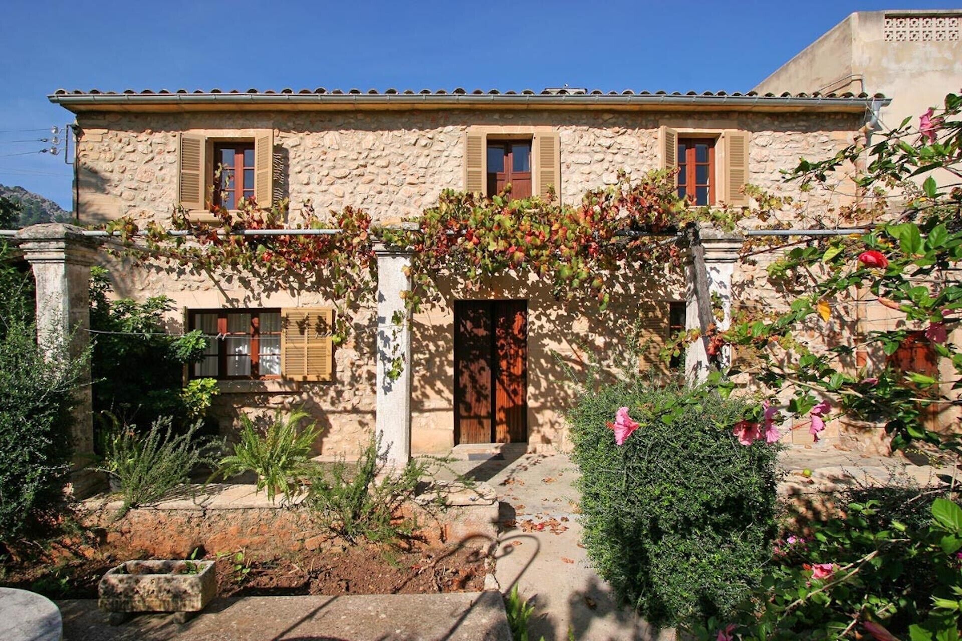 A stone building with a central door, two windows with shutters, and a garden with various plants and a small fountain.