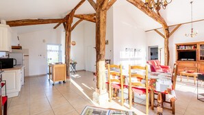 A spacious living area with exposed wooden beams, a dining table with chairs, a sofa, a television, and a kitchen area with a refrigerator and microwave.