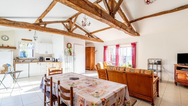 Une cuisine spacieuse avec des meubles blancs, un coin repas agrémenté d’une nappe à motifs floraux et une structure de plafond en bois.