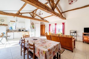 Une cuisine spacieuse avec des meubles blancs, un coin repas agrémenté d’une nappe à motifs floraux et une structure de plafond en bois.