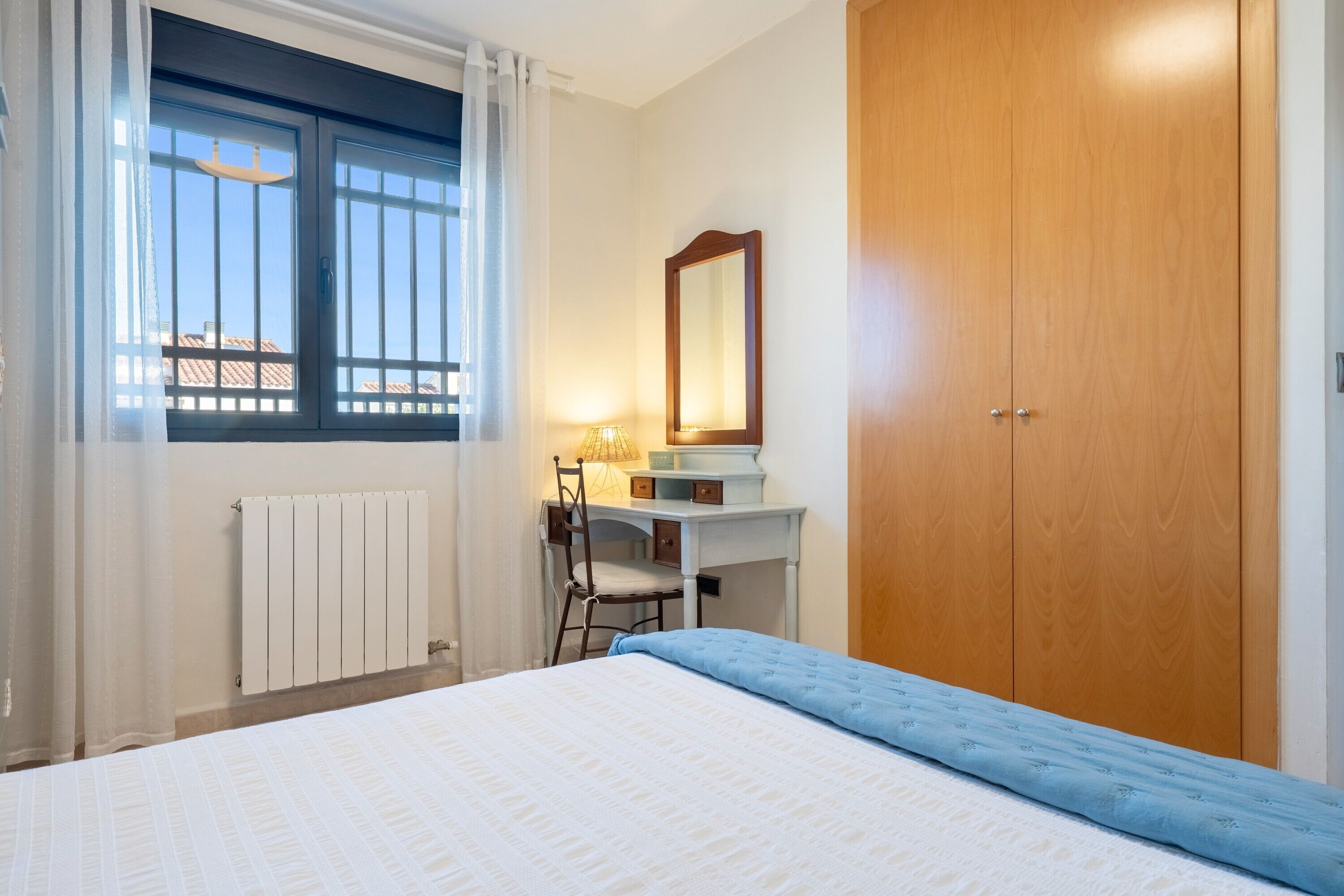 Ein Schlafzimmer mit einem Bett, einem Schreibtisch mit Stuhl, einem Fenster mit Vorhängen, einem Holzschrank und einer Heizung.