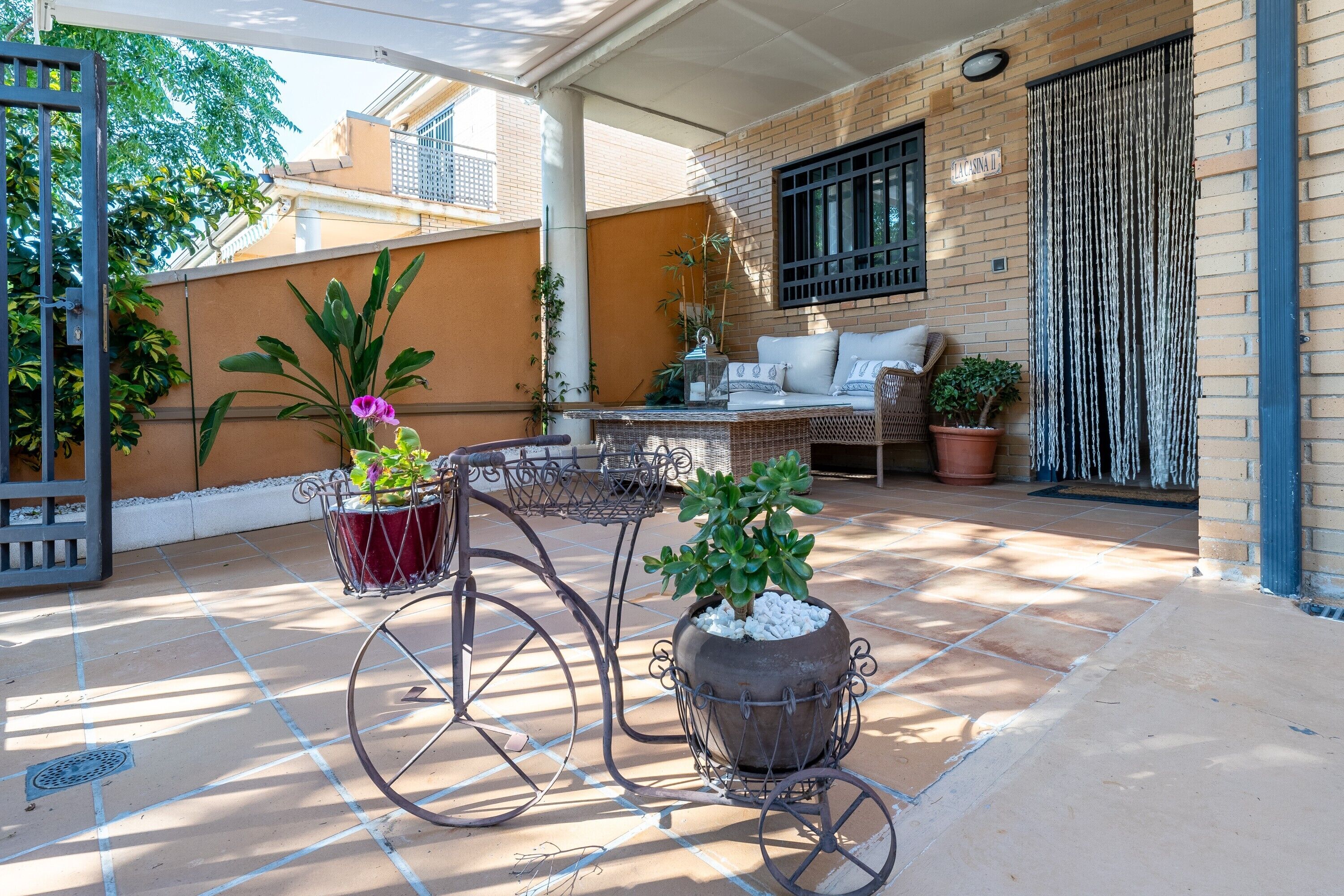 A covered patio with a bicycle planter, a wicker sofa, and a potted plant.