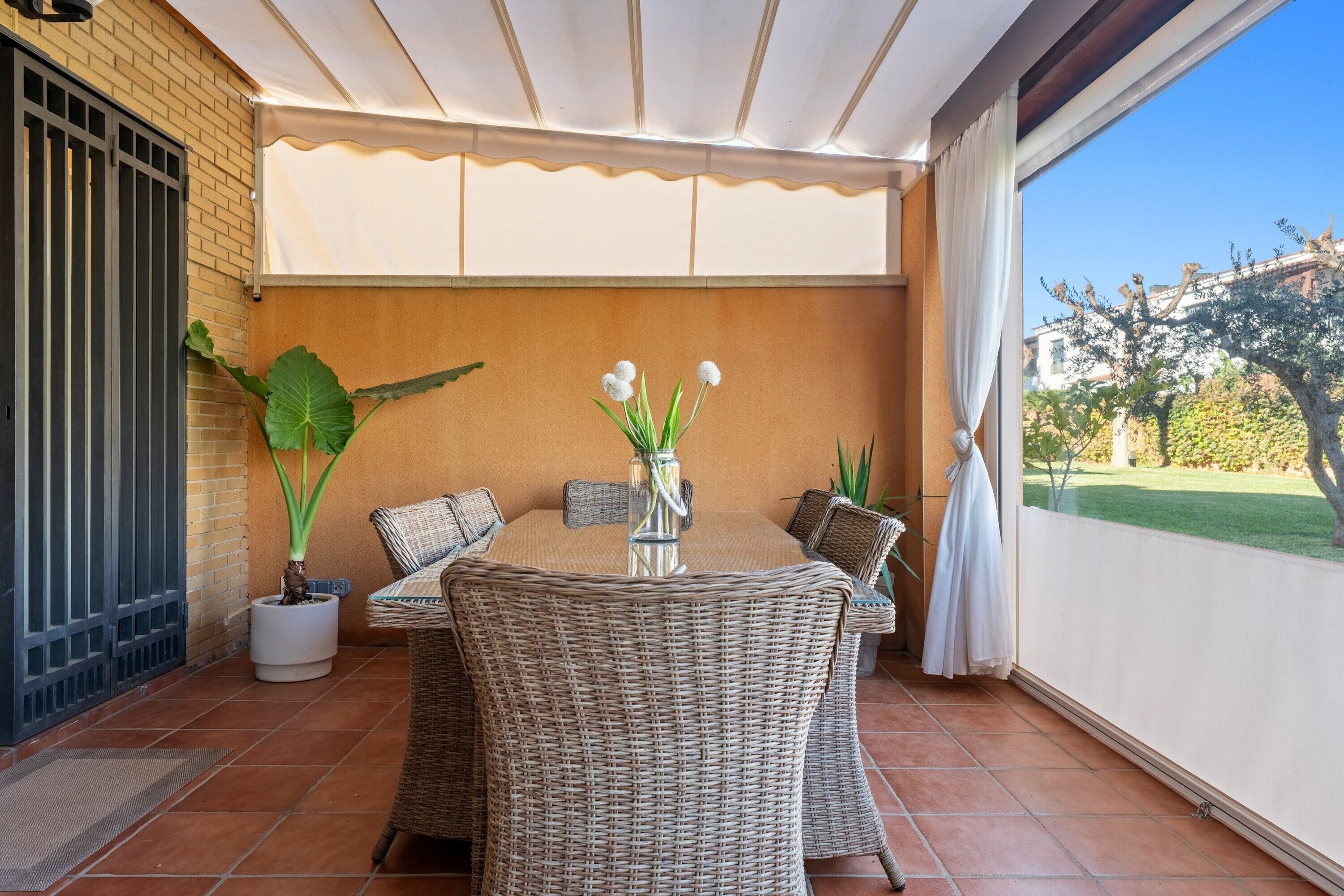 Un patio cubierto con una mesa redonda y sillas de mimbre, una planta en maceta y vistas a un jardín.