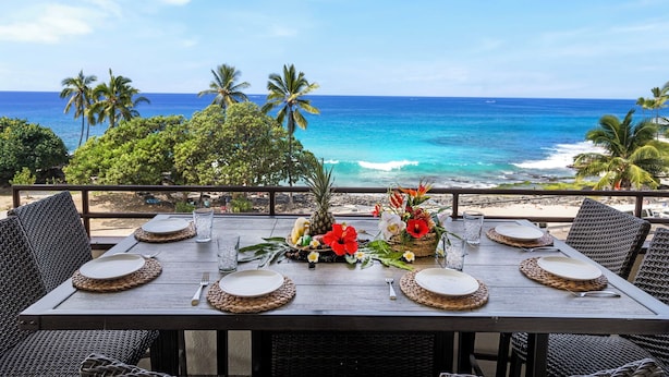 Een eettafel gedekt met borden, bestek en een middelpunt van bloemen en fruit, met uitzicht op een strand met palmbomen en de oceaan.