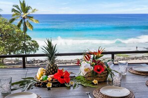 Ein Tisch mit tropischen Früchten, Blumen und Getränken mit Blick auf einen Strand, Wellen und Palmen.