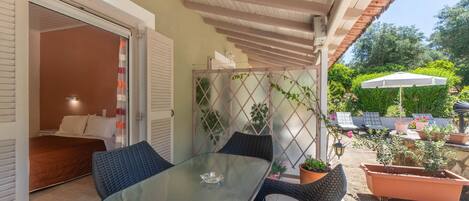 A glass-top table with wicker chairs, a bed with white linens, a potted plant, and a white umbrella in a sunny outdoor area.