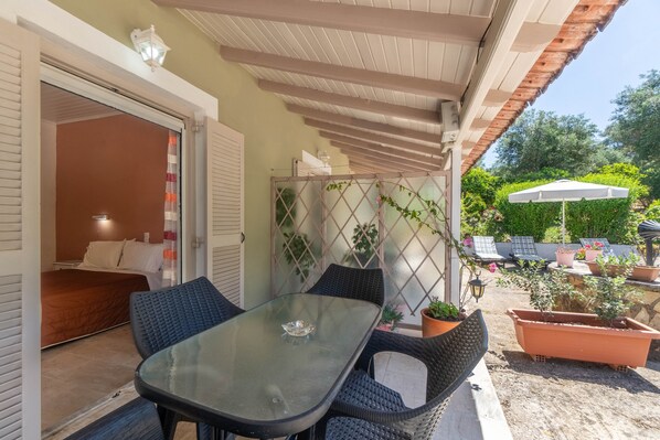 A glass-top table with wicker chairs, a bed with white linens, a potted plant, and a white umbrella in a sunny outdoor area.