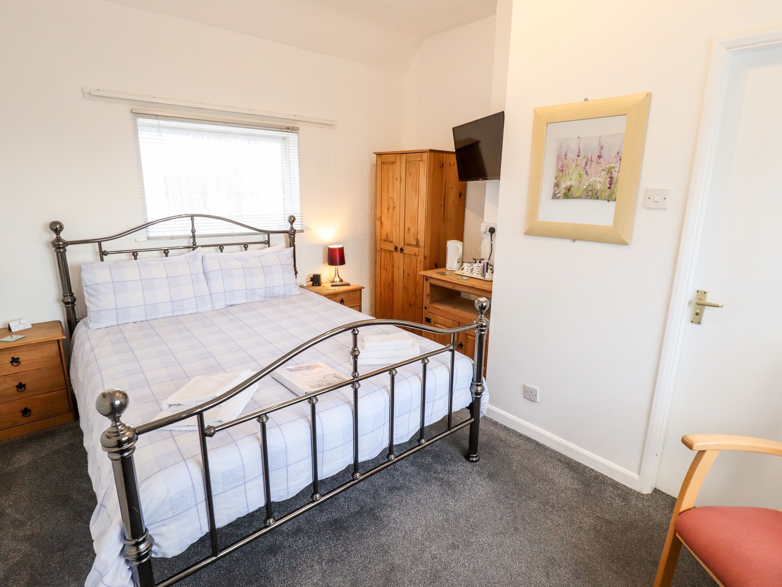 A bedroom with a metal bed frame, a bedside table with a lamp, a wooden wardrobe, a television, and a framed picture on the wall.