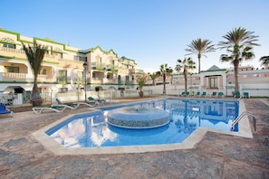Zona de piscina de hotel con un jacuzzi de azulejos, sillas de descanso y palmeras.