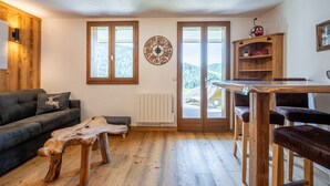 A cozy room with a wooden floor, a sofa with a deer pillow, a wooden coffee table, a clock on the wall, and a view through a door. - Apartment 'Victorsun2' with Mountain View, Terrace and Private Garden (la clusaz)