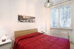 A bedroom with a bed, a nightstand, a lamp, a radiator, and a window with curtains.
