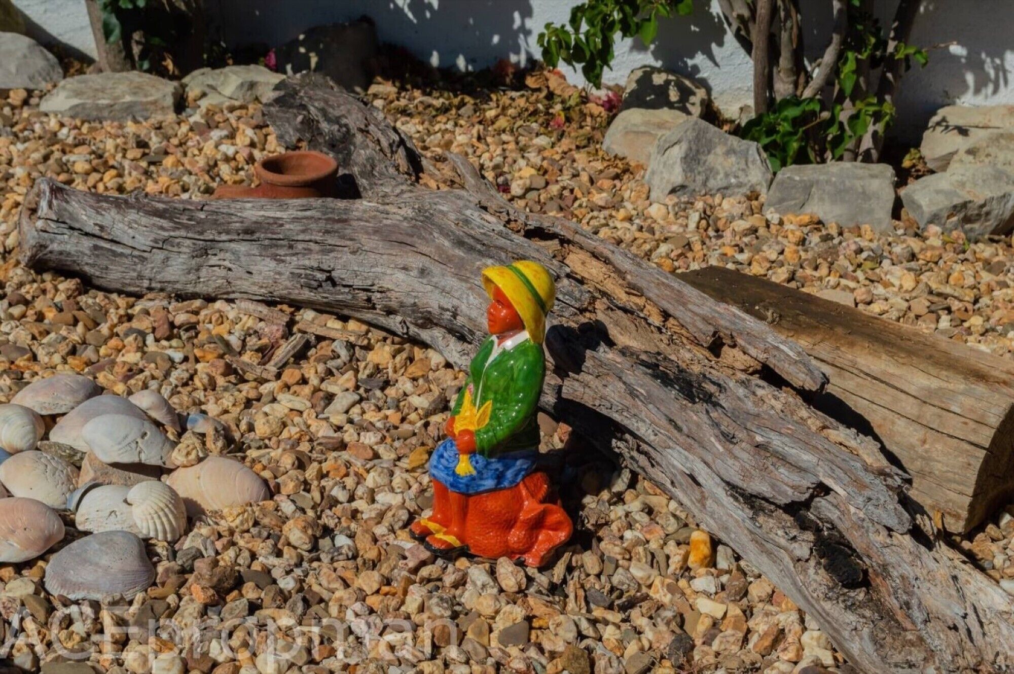 Une petite figurine posée sur une surface de gravier, avec un tronc d’arbre et des rochers en arrière-plan.