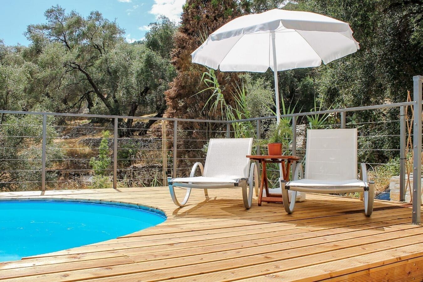 Una terraza de madera con piscina, dos sillas reclinables blancas, una mesa y un paraguas blanco.