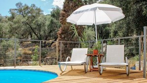 Une terrasse en bois avec une piscine, deux chaises longues blanches, une table et un parasol blanc.