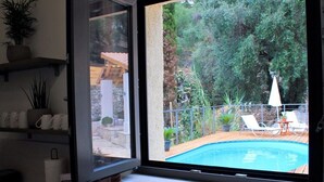 Une fenêtre donnant sur une piscine, du mobilier d’extérieur et de la verdure.