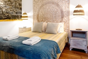 Een net opgemaakt bed met een blauw-geel gestreept dekbed, twee kussens en opgevouwen handdoeken. Aan de linkerzijde een stenen muur en een decoratief hoofdbord met gedetailleerde gravures. Twee wandlampen bevinden zich aan weerszijden van het bed.