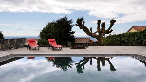 A pool with two red lounge chairs, a stone wall, and a large tree. - Holiday Home 'Ponta Da Amora' with Sea View, Wi-Fi and Air Conditioning (Ponta Garça)