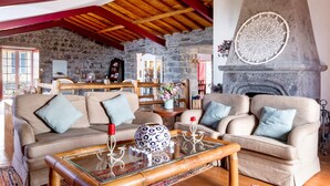 A cozy living room with a stone wall, wooden ceiling beams, a fireplace, and two beige sofas with blue cushions. - Holiday Home 'Ponta Da Amora' with Sea View, Wi-Fi and Air Conditioning (Ponta Garça)