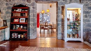 A stone-walled room with a wooden floor, a bookshelf, a dining area with a table and chairs, and a display cabinet with figurines. - Holiday Home 'Ponta Da Amora' with Sea View, Wi-Fi and Air Conditioning (Ponta Garça)