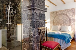 A stone wall, a shower, a wooden chair with a red cushion, and a bed with a blue blanket. - Holiday Home 'Ponta Da Amora' with Sea View, Wi-Fi and Air Conditioning (Ponta Garça)