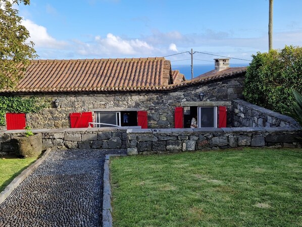 A stone building with a tiled roof, red shutters, and a stone pathway leading to a grassy area. - Holiday Home 'Ponta Da Amora' with Sea View, Wi-Fi and Air Conditioning (Ponta Garça)