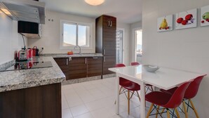 Une cuisine moderne avec des comptoirs en granit, des armoires en bois, une table à manger blanche avec des chaises rouges et une vue sur la ville grâce à de grandes fenêtres.
