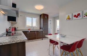 A modern kitchen with granite countertops, wooden cabinets, a white dining table with red chairs, and a view of the city through large windows.