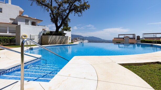Une piscine offrant une vue dégagée sur les montagnes en arrière-plan, un arbre au premier plan et un bâtiment blanc avec un toit recouvert de tuiles.