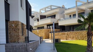 A modern residential building with a concrete pathway leading to a gated entrance, stone walls, and a well-maintained lawn. - Apartment 'Amanecer 7 Alicante' with Shared Pool, Wi-Fi and Air Conditioning (Orihuela)