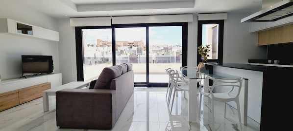 Una sala de estar moderna con un sofá, un televisor y una zona de comedor con sillas y una mesa de vidrio.