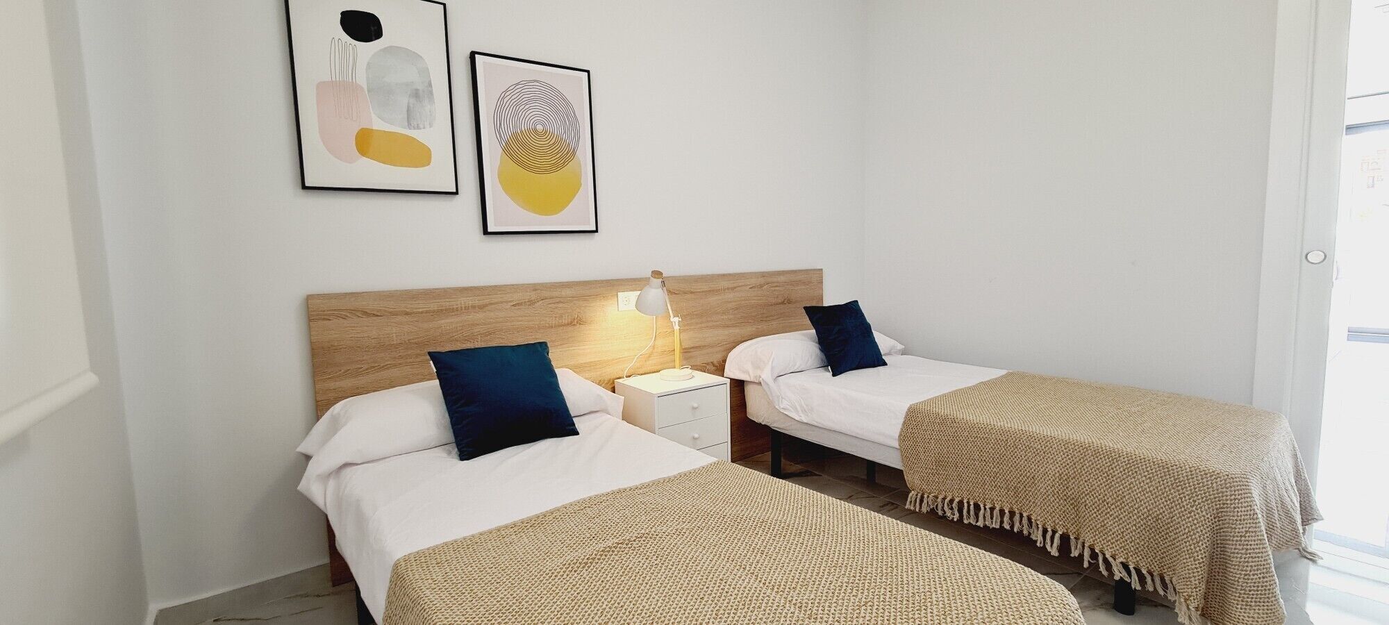 Une chambre d’hôtel avec deux lits, une table de chevet, une lampe et deux œuvres d’art encadrées accrochées au mur.