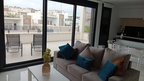 A modern living room with a sofa, a coffee table, and a view of a residential area through large sliding doors. - Apartment 'Amanecer 7 Alicante' with Shared Pool, Wi-Fi and Air Conditioning (Orihuela)