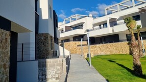 A modern building with a clear sky, a pathway leading to a gated entrance, and a well-maintained lawn. - Apartment 'Amanecer 7 Alicante' with Shared Pool, Wi-Fi and Air Conditioning (Orihuela)