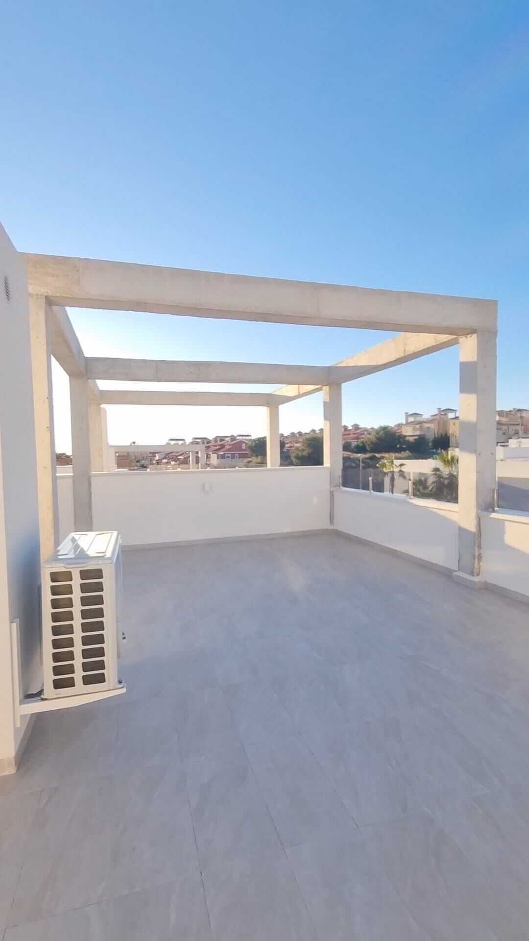 Une terrasse sur le toit, ouverte, avec une structure en béton, un climatiseur blanc et une vue sur les bâtiments environnants.