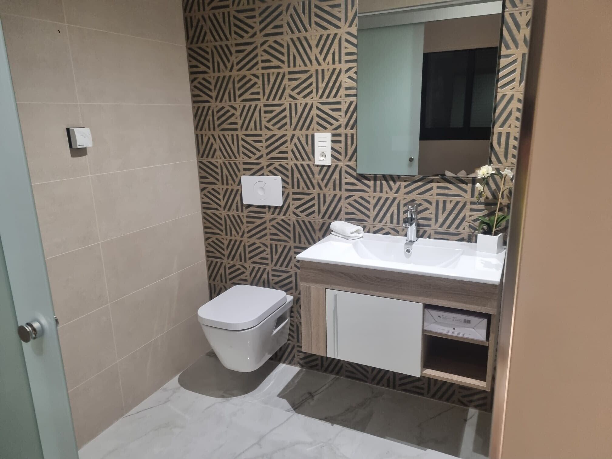 Un baño con pared estampada, inodoro blanco, lavamanos con espejo y un armario.